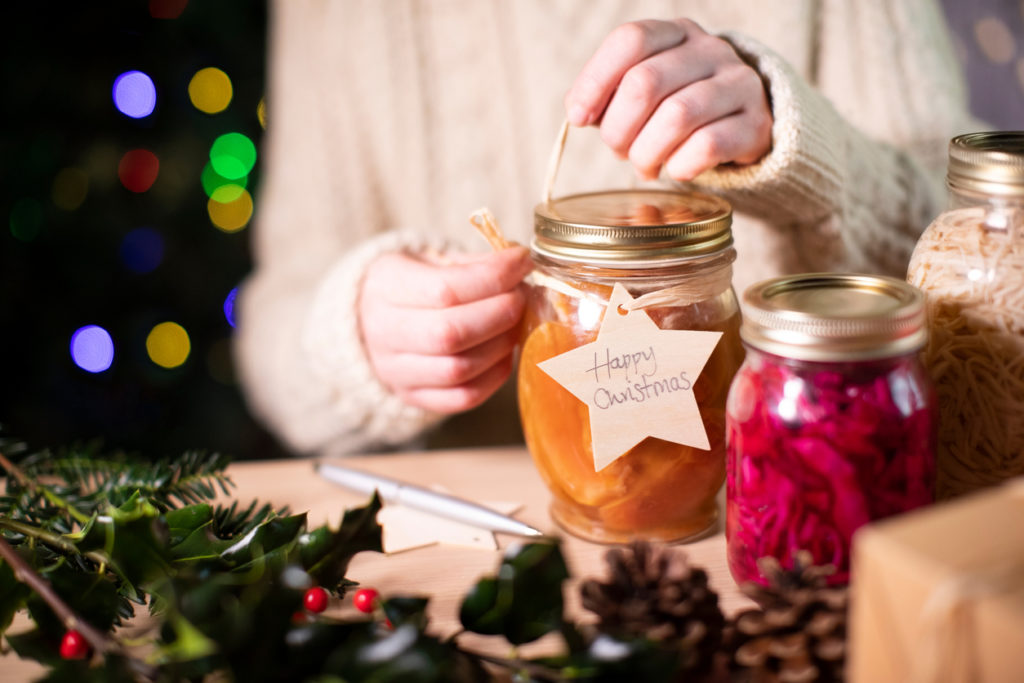 Putting a ribbon and gift tag that says Happy Christmas on a homemade food gift of preserves surrounded by Christmas decor