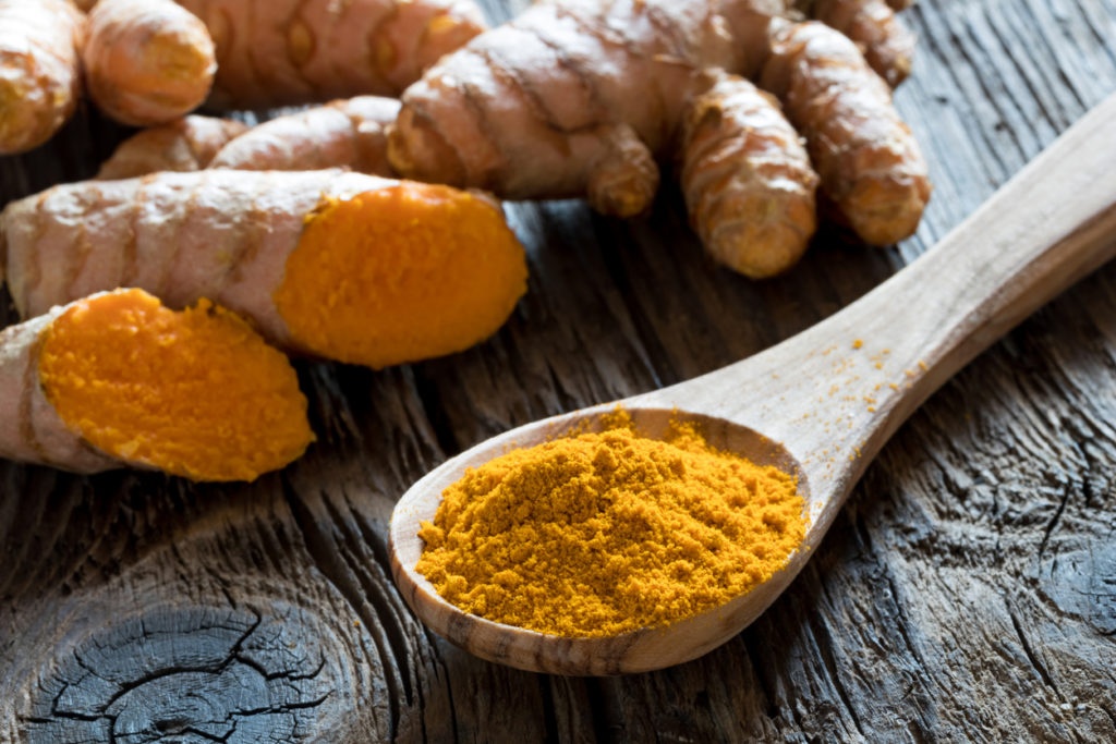 Ground turmeric on a wooden spoon, with fresh turmeric root in the background